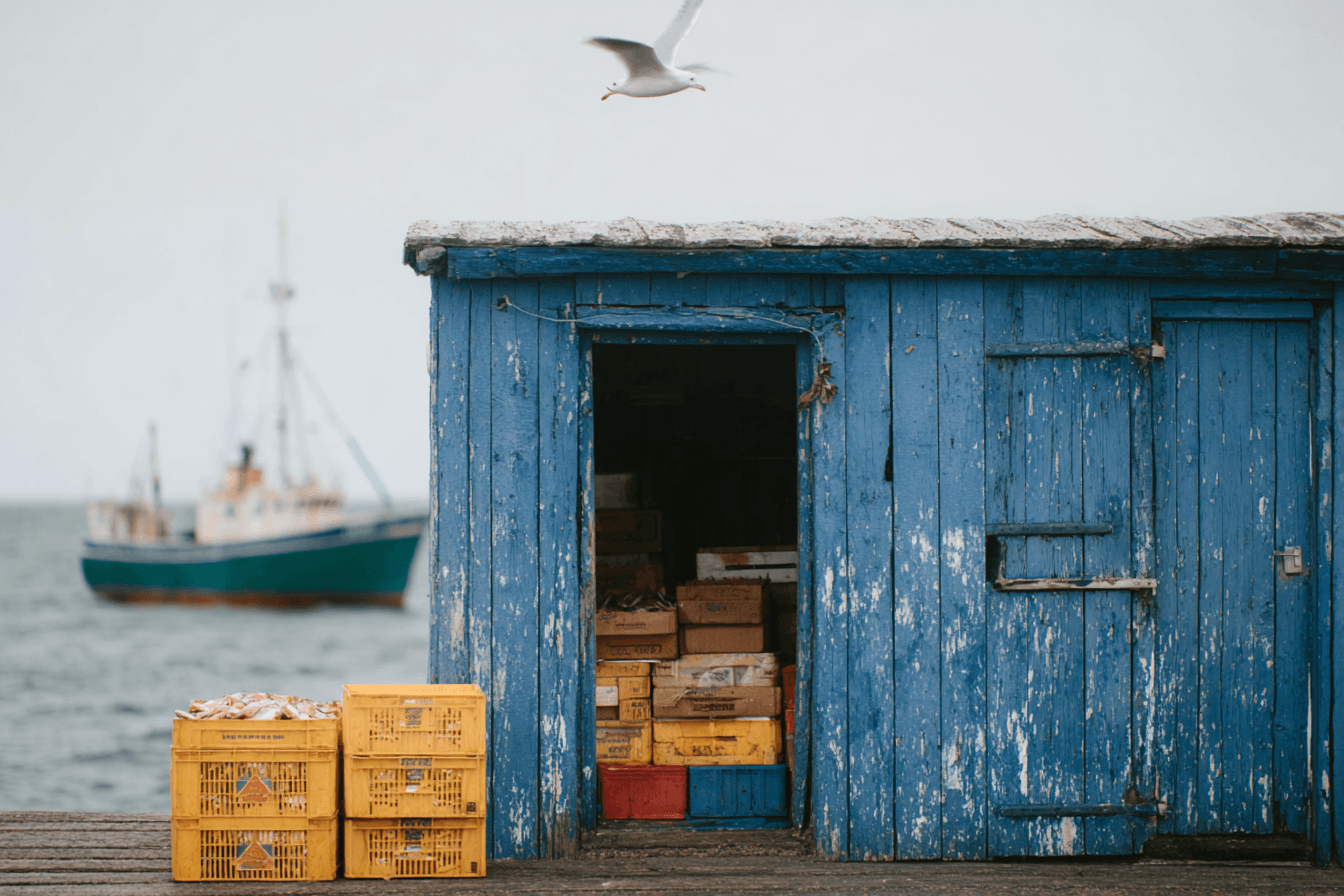 A weathered blue wooden fishing shed on a dock with yellow crates in the foreground and a boat on the water in the background under an overcast sky.